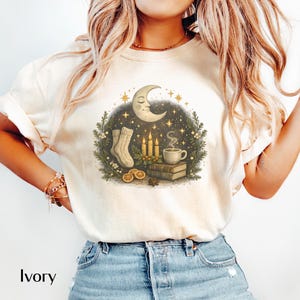 May include: Ivory-colored t-shirt featuring a cozy illustration. The design includes a crescent moon with a smiling face, candles, a cup of coffee, socks, and books. The shirt has a crew neck and short sleeves. The word "Ivory" is printed at the bottom.