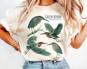 May include: Ivory-colored t-shirt featuring a detailed illustration of green herons in various poses. The design includes the text "GREEN HERON" and "Butorides virescens". The shirt is made of a soft material and is suitable for casual wear.