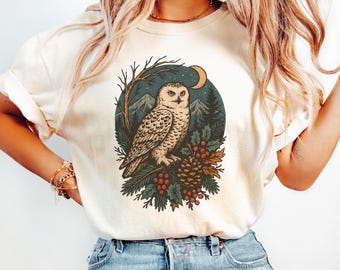 May include: Ivory-colored t-shirt featuring a detailed illustration of a snowy owl perched on a branch, surrounded by pine needles, berries, and a pinecone. The design includes a crescent moon and a backdrop of mountains and trees. The word "Ivory" is printed at the bottom.