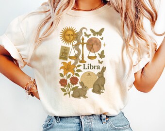 Op de afbeelding: Ivoor kleurig T-shirt met een Weegschaal sterrenbeeld ontwerp. De afbeelding bevat een zon, weegschalen, vlinders, konijnen, bloemen en het woord "Weegschaal". Het shirt is gemaakt van een zacht materiaal.