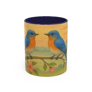 May include: A blue and orange ceramic mug with a blue rim. The mug features a watercolor illustration of two bluebirds perched on a branch with pink blossoms.