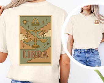 Op de afbeelding: Ivoor kleurig T-shirt met een Weegschaal sterrenbeeld ontwerp. Het ontwerp bevat een weegschaal, sterren en wolken in bruin, groen en goudtinten. Het woord "LIBRA" staat eronder gedrukt. Een kleiner Weegschaal symbool staat op de voorkant.