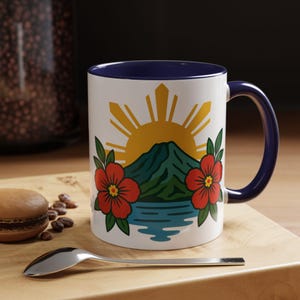 May include: White ceramic mug with a blue interior and handle. The mug features a design of a yellow sun, green mountain, blue water, and red flowers. A spoon and a pastry are in the foreground.