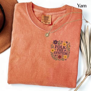 May include: A coral-colored t-shirt with a crew neck and the words "FOLK THE SYSTEM" in a floral design. A gold necklace with a pendant is draped across the shirt. The shirt has a Comfort Colors tag.