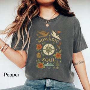 May include: Pepper-colored t-shirt with a graphic design featuring a compass rose, birds, flowers, and the words "Nomadic Soul." The design includes a backpack, footprints, and a mountain scene. The shirt is a relaxed fit.