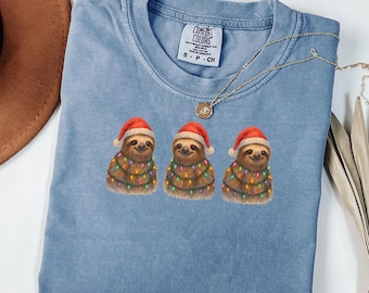 May include: A blue jean colored t-shirt featuring three sloths wearing Santa hats and Christmas lights. The shirt has a Comfort Colors tag and a gold necklace. The text "Blue Jean" is at the top.