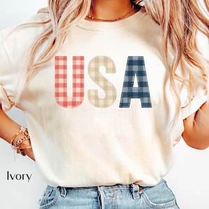 Peut inclure: T-shirt ivoire avec le mot "USA" dans un motif à carreaux. Les lettres sont rouge/rose, beige et bleu/marine. Le mot "Ivory" est imprimé en bas du t-shirt.