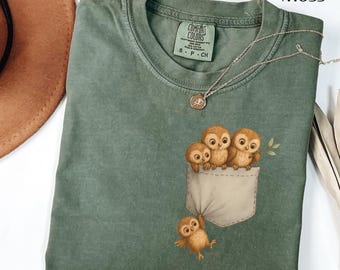 May include: A moss green t-shirt featuring a pocket design with three owls peeking out and one hanging below. The shirt has a crew neck and a small tag. A silver necklace with a pendant is draped across the shirt.