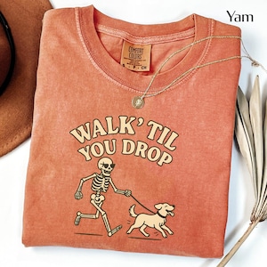 May include: A yam-colored t-shirt with the phrase "WALK 'TIL YOU DROP" printed on the front. The graphic shows a skeleton wearing sunglasses walking a dog. A gold necklace is draped across the shirt.