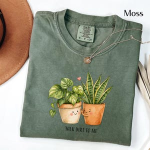May include: Moss green t-shirt with a graphic of two potted plants with smiling faces and the text "TALK DIRT TO ME". A silver necklace with a small pendant is draped across the shirt.