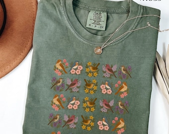 May include: A moss green t-shirt with a crew neck and short sleeves. The shirt features a repeating pattern of colorful birds and flowers. A silver necklace with a small pendant rests on the shirt.