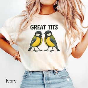 May include: Ivory-colored t-shirt featuring the text "GREAT TITS" above a graphic of two yellow and black birds. The birds are facing each other. The shirt is made of a soft material.