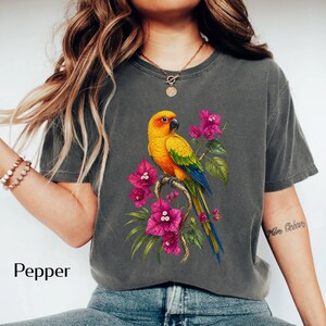 May include: Dark gray t-shirt featuring a vibrant illustration of a yellow, orange, and green parrot perched on a branch with pink flowers. The word "Pepper" is printed in white at the bottom left.