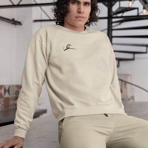 May include: A person wearing a light beige sweatshirt with a small embroidered bird design on the left chest. The person is sitting on a concrete surface.
