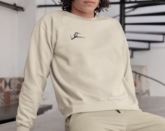 May include: A person wearing a light beige sweatshirt with a small embroidered bird design on the left chest. The person is sitting on a concrete surface.