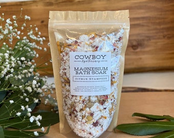 Könnte beinhalten: Ein transparenter Beutel gefüllt mit Badesalz. Das Etikett lautet "Cowboy Apothecary Magnesium Bath Soak Citrus Stampede". Das Bad enthält weiße Granulate, getrocknete Blütenblätter und andere natürliche Inhaltsstoffe. Der Hintergrund ist Holz.