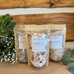 May include: Three clear pouches of Cowboy Apothecary Magnesium Bath Soak. The pouches are labelled with the scents: Lavender, Citrus Stampede, and Peppermint. The bath soaks contain salts and botanicals. Greenery and wood planks are in the background.