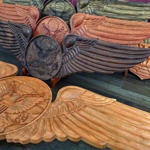 May include: A collection of intricately carved wooden eagle crests and wings in various shades of brown and reddish-brown. The carvings feature detailed feather textures and the United States seal, showcasing craftsmanship and patriotic themes.