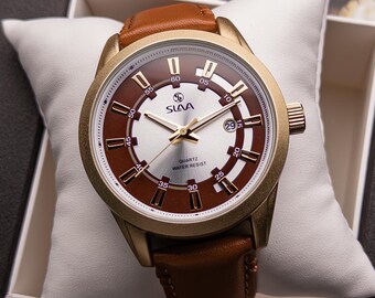 May include: A gold-tone wristwatch with a brown leather strap. The watch face has a white background with brown and gold accents. The watch face also has the text "SLAVA QUARTZ WATER RESIST" printed on it.