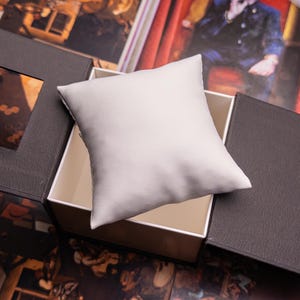 May include: A black and white box with a white cushion inside. The box is open and the cushion is visible. The box is made of a textured material.