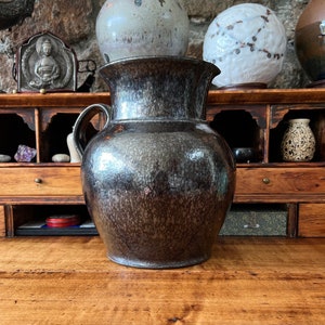 May include: A dark brown ceramic pitcher with a speckled glaze. The pitcher has a wide mouth and a handle. It is sitting on a wooden table.