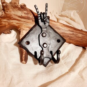 Könnte beinhalten: Schwarzer Wandhaken aus Metall in Form einer Hand, die das Rock'n'Roll-Handzeichen macht. Der Haken hat zwei Zacken zum Aufhängen von Gegenständen. Der Haken ist auf einer quadratischen Metallplatte montiert.