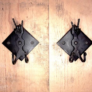 May include: Two black metal wall hooks with a diamond-shaped base. Each hook features a hand-forged design with a peace sign gesture.