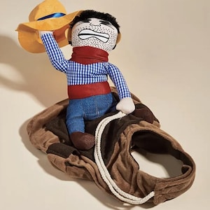 May include: A brown plush horse costume for pets, featuring a cowboy doll riding on top. The cowboy doll is wearing a blue and white plaid shirt, red vest, blue jeans, and a yellow cowboy hat. The doll has a cartoon face with a surprised expression.