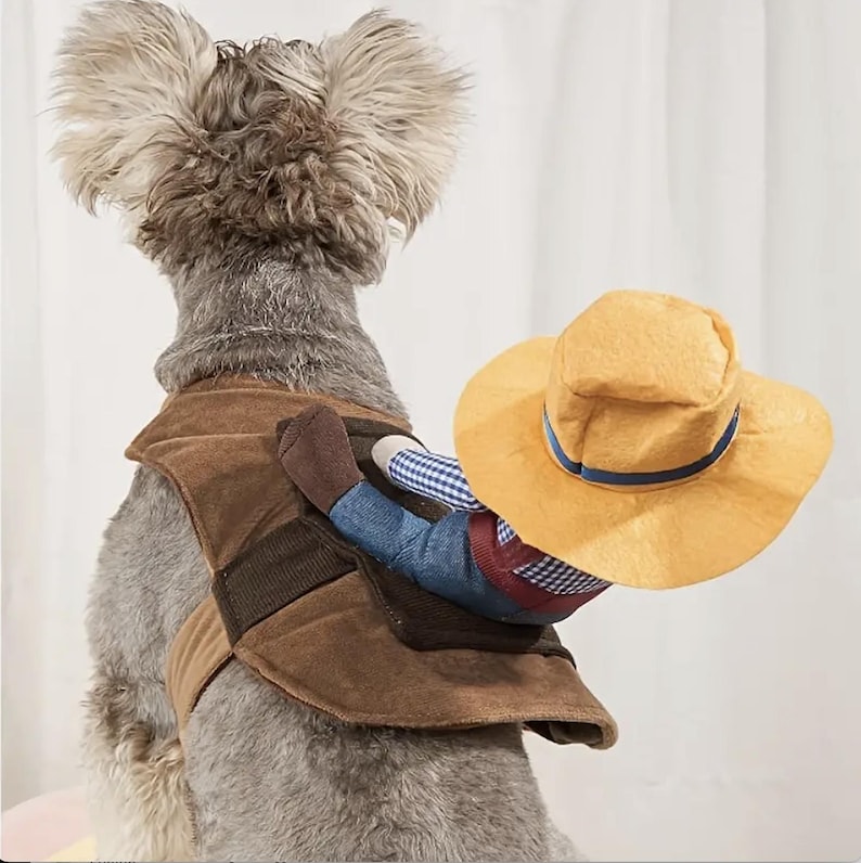 May include: A brown and tan dog costume features a plush cowboy riding on the dog's back. The cowboy is wearing a blue and white plaid shirt, blue jeans, brown boots, and a yellow straw cowboy hat with a blue band.