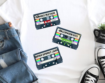 Puede incluir: Camiseta blanca con tres diseños de cintas de cassette. Las cintas tienen detalles de arcoíris y los textos "Spooky Mixtape", "Halloween Hits Vol 1" y "Retro Spooky Jams". La camiseta se muestra con vaqueros azules y zapatillas negras.