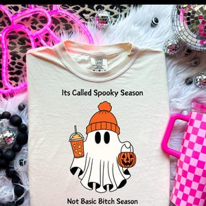 Könnte beinhalten: Ein weißes T-Shirt mit einem Cartoon-Geist, der eine orangefarbene Mütze trägt und einen Kürbis und einen Kürbisgewürz-Latte hält. Der Text auf dem Shirt lautet "Its Called Spooky Season Not Basic Bitch Season".
