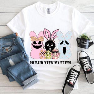 Puede incluir: Camiseta blanca con un gráfico de tres peeps de dibujos animados sosteniendo huevos de Pascua. El texto "Chillin with my peeps" está impreso debajo del gráfico.