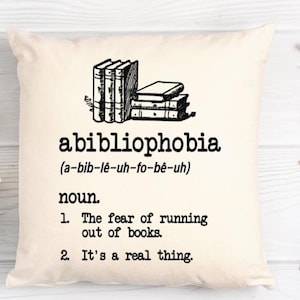 May include: A cream-coloured square cushion featuring the word "abibliophobia" and its pronunciation in black text. Illustrations of books are above the text. The definition of abibliophobia is also printed on the cushion.