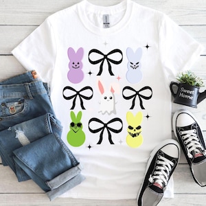 Può includere: Maglietta bianca con una grafica di peeps pasquali color pastello con fiocchi neri. I peeps hanno diverse espressioni facciali, tra cui un fantasma, un teschio e occhiali da sole.
