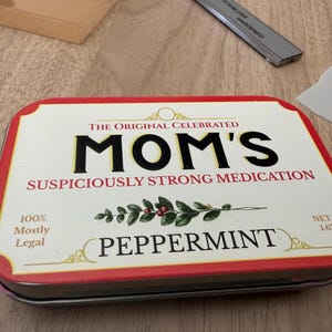 May include: A rectangular metal tin with a red border and a white label. The label reads "MOM'S SUSPICIOUSLY STRONG MEDICATION PEPPERMINT." The tin also states "100% Mostly Legal" and "NET WT 1.67 OZ."