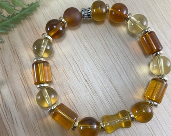 May include: A beaded bracelet featuring a mix of amber and yellow-toned beads. The bracelet includes round, cylindrical, and barrel-shaped beads, separated by small gold-colored spacers. The beads are strung on an elastic band.