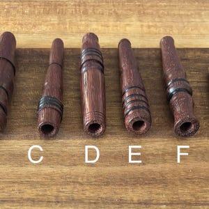 May include: A collection of eight wooden smoking pipes in various designs. The pipes are primarily brown with black accents, and some feature striped patterns. The pipes are arranged on a wooden surface, labeled A through H.