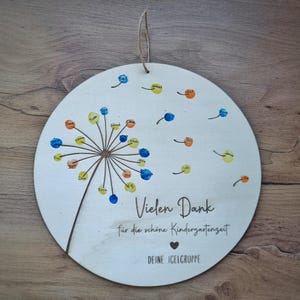 Könnte beinhalten: Ein weißer Holz-Kreis mit einem gemalten Löwenzahnmuster. Der Löwenzahn hat blaue, gelbe und orange Fingerabdrücke. Der Text "Vielen Dank für die schöne Kindergartenzeit" und "DEINE IGELGRUPPE" steht auf dem Kreis.