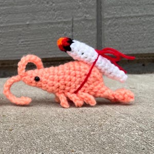 May include: A handmade crochet shrimp in a light coral color, with a white and red crochet cigarette attached to its back. The shrimp has black eyes and red yarn accents. The shrimp is resting on a concrete surface.