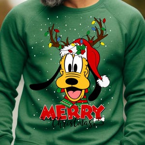 Może przedstawiać: Zielony świąteczny sweter z kreskówkowym wizerunkiem Plutona w czapce Świętego Mikołaja i rogami renifera. Sweter ma napis "Merry Christmas".