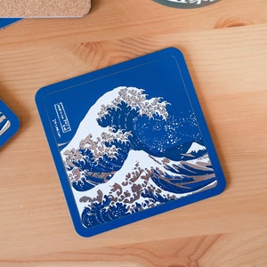 May include: Square blue coaster featuring a print of Hokusai's "The Great Wave off Kanagawa." The artwork depicts a large ocean wave with a mountain in the background. The coaster has a dark blue border and a light wood grain background.