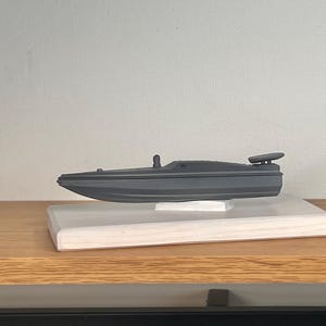 以下が含まれることがあります： 小さなアンテナとフィンが付いたグレーの模型のスピードボート。白い木の台に展示されています。ボートは、ニュートラルな壁を背景に、木の棚に置かれています。