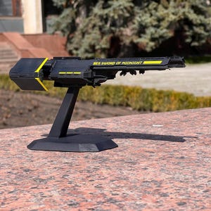 May include: A black spaceship model with yellow detailing, mounted on a black stand. The text "SES SWORD OF MIDNIGHT" is visible on the side. The model is displayed outdoors on a stone surface.