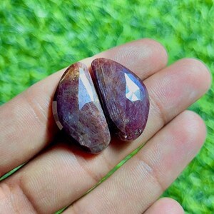 Puede incluir: Dos piedras preciosas facetadas en forma de lágrima con un tono rojo oscuro y púrpura. Las piedras presentan una superficie pulida y reflectante con inclusiones internas visibles y un borde recto y plano en un lado, aspecto semiprecioso.