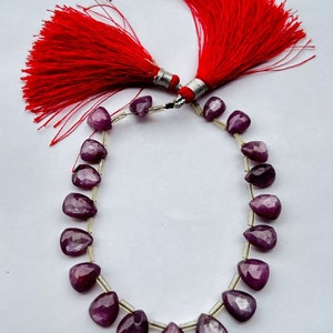 Puede incluir: Un collar de piedras preciosas de color rojo púrpura intenso, posiblemente rubíes, con forma de lágrima, ensartadas en un fino alambre plateado. Dos borlas rojas brillantes con detalles plateados en la parte superior de la imagen.