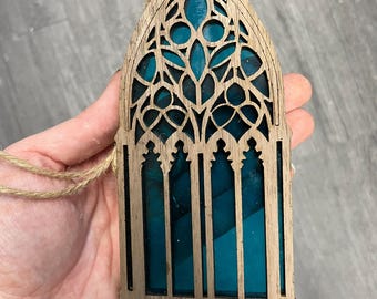 Könnte beinhalten: Hölzernes gotisches Fensterornament mit türkisblauem Buntglas. Das Ornament hat ein detailliertes, durchbrochenes Design und wird von einer natürlichen Schnurschlaufe gehalten. Der Fensterrahmen besteht aus hellbraunem Holz.