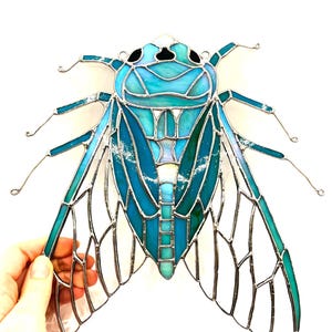 May include: A stained glass cicada in shades of teal and turquoise, with silver wire detailing. The insect's wings are translucent, and the body is segmented. The artwork is held against a white background.