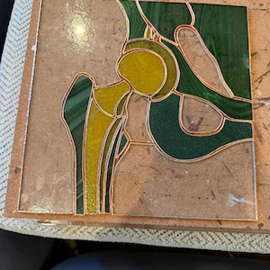 Op de afbeelding: Gebrandschilderd glaspaneel met een menselijk heupgewricht. Het kunstwerk toont een combinatie van geel en groen glas, omlijnd met koperfolie. Het ontwerp is geplaatst tegen een neutrale achtergrond en toont de anatomische vorm.
