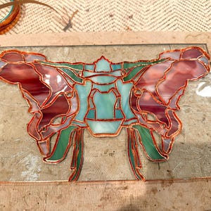 May include: A stained glass piece in progress, featuring a butterfly design. The artwork uses red, pink, green, and blue glass, outlined with copper foil. The glass is set on a textured, light brown surface.