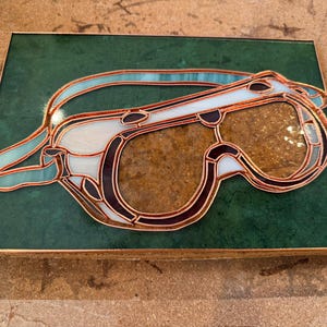 May include: Stained glass artwork depicting a pair of goggles. The goggles feature a copper frame with clear, brown, white, and teal glass pieces. The artwork is set on a green background and framed in a wooden border.